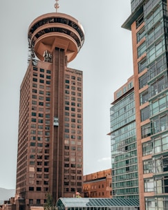 A tall tower with a unique circular observation deck at the top, surrounded by modern glass buildings. The architecture features numerous windows and a rooftop antenna.