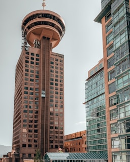 A tall tower with a unique circular observation deck at the top, surrounded by modern glass buildings. The architecture features numerous windows and a rooftop antenna.