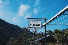 A scenic view of a signpost with Japanese characters, against a backdrop of a clear blue sky and a mountainous landscape. The sign is attached to a metallic structure, and there is lush greenery surrounding the area.