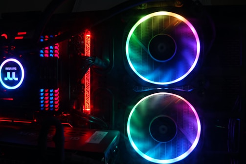 A computer setup featuring vibrant RGB lighting with two large cooling fans illuminated in a spectrum of colors. Cables and components are visible, including a power supply and various heat sinks. The lighting creates a futuristic and dynamic appearance.