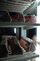 Several large cuts of raw beef are arranged on metal racks inside a refrigerator. The meat appears to be aging, with a dark, crusty exterior and rich marbling. The refrigerator is illuminated by bright lights, accentuating the textures and colors of the meat.