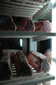 Several large cuts of raw beef are arranged on metal racks inside a refrigerator. The meat appears to be aging, with a dark, crusty exterior and rich marbling. The refrigerator is illuminated by bright lights, accentuating the textures and colors of the meat.