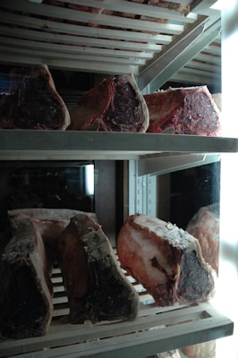 Several large cuts of raw beef are arranged on metal racks inside a refrigerator. The meat appears to be aging, with a dark, crusty exterior and rich marbling. The refrigerator is illuminated by bright lights, accentuating the textures and colors of the meat.