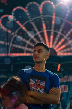 A young man stands confidently with his arms crossed, wearing a blue shirt featuring a Union Jack and the phrase 'Keep Calm and Listen to Music.' Behind him, a light display with circular patterns glows in various colors, creating a festive atmosphere. A child with reddish-brown hair in a pink shirt is situated in the foreground.