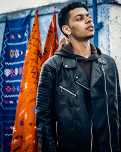 A young man wearing a stylish embroidered jacket with Arabic calligraphy, standing against a vibrant city mural.