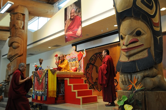 A ceremonial event featuring several monks dressed in traditional robes. They are gathered around an ornately decorated stage with religious artifacts. The setting includes large, intricately carved wooden totem poles and a photograph of a smiling figure displayed prominently.