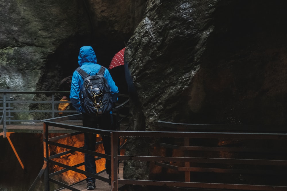 Persona con chaqueta azul con capucha de pie en un puente peatonal cerca de la cueva