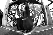 A person stands in front of a wall covered with abstract black and white graffiti, holding a large bag. The wall features swirling, dynamic patterns. The scene gives a sense of motion and urban art culture.