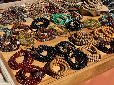 A variety of beaded bracelets are displayed on a wooden tray. The bracelets are made up of beads in different colors and materials, including browns, greens, blacks, and some mixed with turquoise and gold tones. The beads vary in size and are arranged in a way to showcase the diversity of designs and colors.
