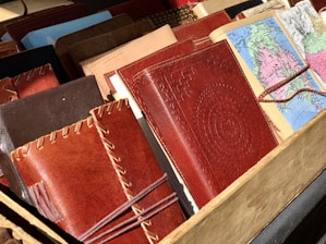 A collection of various leather-bound notebooks and journals, some featuring intricate stitching and embossed patterns. Next to them, there are maps folded into book-sized brochures. They are arranged in a wooden display.