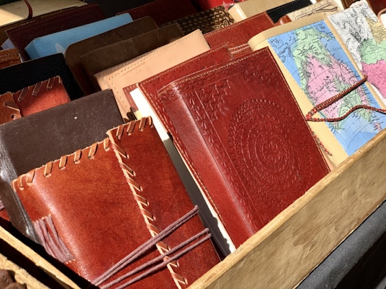 A collection of various leather-bound notebooks and journals, some featuring intricate stitching and embossed patterns. Next to them, there are maps folded into book-sized brochures. They are arranged in a wooden display.