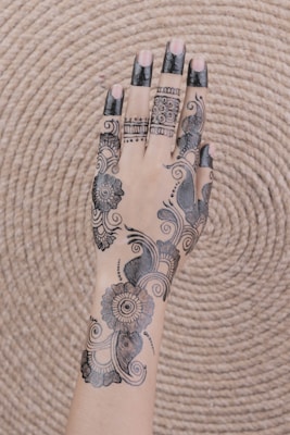 A hand adorned with intricate henna designs, featuring floral patterns and swirls, with sections of the fingers coated in black henna. The background is a woven, circular mat that provides a textured contrast to the skin and artwork.