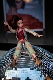 A doll with brown hair is sitting on top of a reflective disco ball. The doll is dressed in a stylish outfit with a red top, beige jacket, and green pants. It has boots on its feet and appears to be waving.