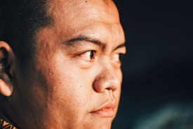 A close-up of a person's face with a focused expression, highlighting their features and skin texture. The lighting creates shadows and emphasizes the details of the skin, giving a dramatic effect.