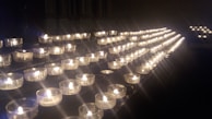 A series of small tea light candles arranged in rows on a dark surface, each held in a clear glass container. The candles emit a warm, soft glow, creating an atmosphere of tranquility and contemplation.