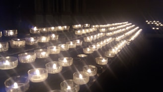 A series of small tea light candles arranged in rows on a dark surface, each held in a clear glass container. The candles emit a warm, soft glow, creating an atmosphere of tranquility and contemplation.