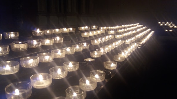 A series of small tea light candles arranged in rows on a dark surface, each held in a clear glass container. The candles emit a warm, soft glow, creating an atmosphere of tranquility and contemplation.