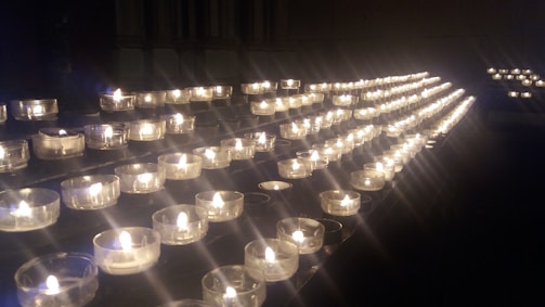 A series of small tea light candles arranged in rows on a dark surface, each held in a clear glass container. The candles emit a warm, soft glow, creating an atmosphere of tranquility and contemplation.