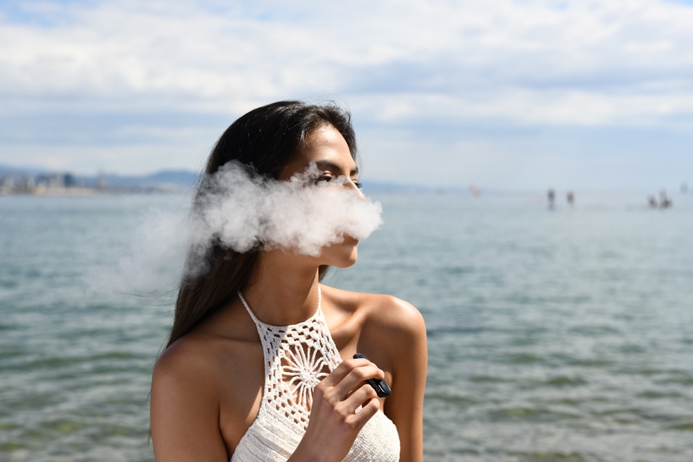 woman wearing white sleeveless top smoking tobacco while standing near blue sea under white and blue skies during daytime