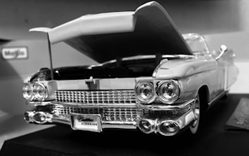 A vintage car model with an open hood, showcasing intricate details of the front grille, headlights, and bumper. The car appears to be a detailed replica with a classic design, featuring the Maisto brand label in the background.