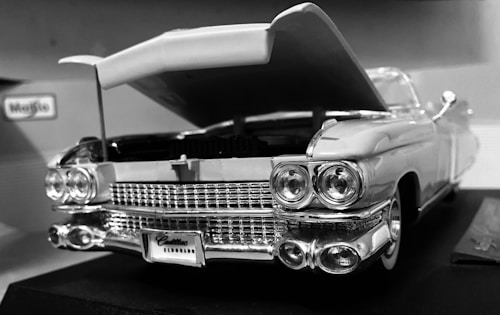 A vintage car model with an open hood, showcasing intricate details of the front grille, headlights, and bumper. The car appears to be a detailed replica with a classic design, featuring the Maisto brand label in the background.