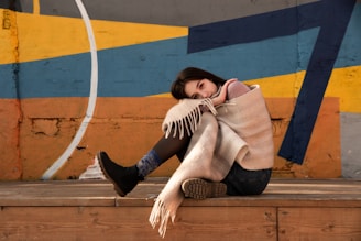 A person is sitting on a wooden stage or platform, wrapped in a beige blanket. The individual is resting their head on their knee, with a contemplative expression. The background features a colorful, abstract mural with geometric shapes in shades of yellow, blue, and orange. The person's attire includes black boots and denim jeans.