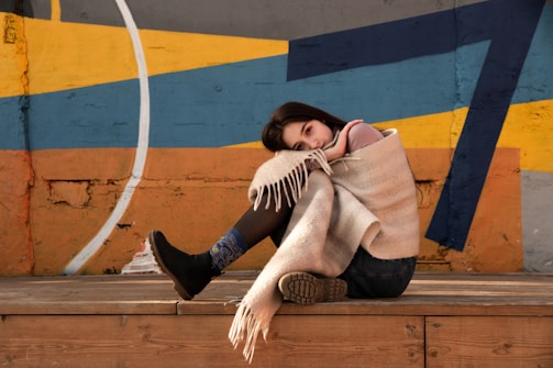 A person is sitting on a wooden stage or platform, wrapped in a beige blanket. The individual is resting their head on their knee, with a contemplative expression. The background features a colorful, abstract mural with geometric shapes in shades of yellow, blue, and orange. The person's attire includes black boots and denim jeans.