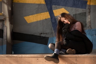A person with long hair sits on a wooden platform, leaning slightly with a contemplative expression. She is wearing dark clothing and sneakers. The background is an abstract mural with blue, yellow, and gray geometric patterns that suggests an urban environment.