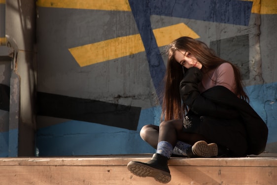 A person with long hair sits on a wooden platform, leaning slightly with a contemplative expression. She is wearing dark clothing and sneakers. The background is an abstract mural with blue, yellow, and gray geometric patterns that suggests an urban environment.