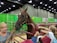 A horse is standing inside a red metal enclosure with green curtains in the background. Several people, including children and an adult, are reaching out to touch the horse. The surroundings suggest an indoor event or exhibition space.
