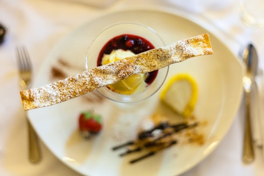A decorative dessert is elegantly presented on a white plate, featuring a glass filled with a dessert topped with a red berry sauce and a slice of ice cream. A crispy pastry stick, dusted with powdered sugar, is placed across the top. Garnishes include a fresh strawberry and a lemon slice on the side. The setup is surrounded by cutlery, indicating a fine dining ambiance.