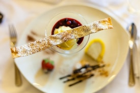 A decorative dessert is elegantly presented on a white plate, featuring a glass filled with a dessert topped with a red berry sauce and a slice of ice cream. A crispy pastry stick, dusted with powdered sugar, is placed across the top. Garnishes include a fresh strawberry and a lemon slice on the side. The setup is surrounded by cutlery, indicating a fine dining ambiance.
