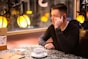 A person sitting at a table in a cozy indoor setting, holding a phone to their ear. They are next to a window with warm decorative lighting. A cup and saucer, a bowl, and a menu are also on the table, suggesting a cafe or restaurant environment.