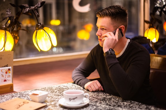 A person sitting at a table in a cozy indoor setting, holding a phone to their ear. They are next to a window with warm decorative lighting. A cup and saucer, a bowl, and a menu are also on the table, suggesting a cafe or restaurant environment.