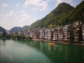A serene river flows through a town with traditional multi-story buildings on its banks, characterized by a mix of modern and classic architecture. Lush green hills rise in the background under a partly cloudy sky, creating a picturesque landscape. A single small boat with a red canopy glides across the calm waters of the river.