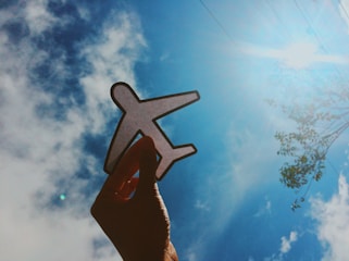 A hand is holding a paper cutout of an airplane against a bright blue sky with scattered clouds. Sunlight shines from the top right corner, creating a vibrant and optimistic atmosphere.