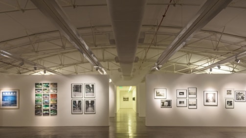 An art gallery with white walls and a high ceiling features various framed photographs and artworks displayed in a well-organized arrangement. The room is brightly lit, emphasizing the artwork. The ceiling has exposed beams and lighting fixtures.
