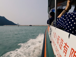 A boat is moving across a body of water, with splashing waves visible near its side. Several people are onboard, and a mountain can be seen in the background. There is a sign on the boat with text in both English and another language. A breakwater and navigational marker are in the distance.