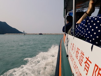 A boat is moving across a body of water, with splashing waves visible near its side. Several people are onboard, and a mountain can be seen in the background. There is a sign on the boat with text in both English and another language. A breakwater and navigational marker are in the distance.