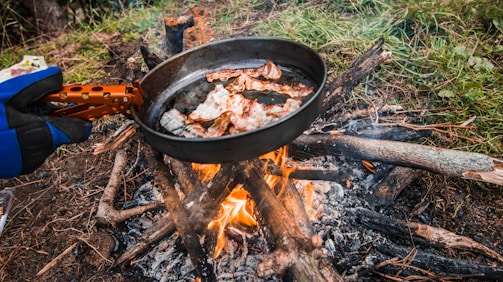 A person wearing a glove holds a frying pan filled with strips of bacon over a campfire made from wooden logs. The flames lick the bottom of the pan, and the scene is set outdoors on a grassy area surrounded by green vegetation.