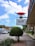 A street scene with a prominent sign featuring a pair of red lips above a message that reads 'ALL WAYS WELCOME'. The setting includes a sidewalk lined with plants and a row of shops or businesses. Several cars are on the street, and a clear blue sky with some clouds is visible above.