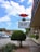 A street scene with a prominent sign featuring a pair of red lips above a message that reads 'ALL WAYS WELCOME'. The setting includes a sidewalk lined with plants and a row of shops or businesses. Several cars are on the street, and a clear blue sky with some clouds is visible above.