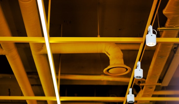 A ceiling with industrial elements, featuring exposed pipes and beams painted in a bright yellow color. Several cylindrical track lights are mounted on the beams, providing illumination. The overall feel is modern and industrial.