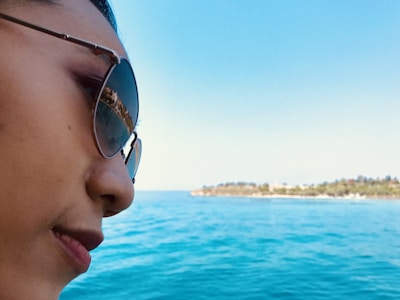 Stylish individual enjoying outdoor sunlight wearing sunglasses with a serene beach background.