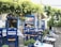 A charming outdoor restaurant area with blue wooden chairs and white tables, set in a quaint stone courtyard. Green vines and plants create a natural canopy overhead, providing shade and a cozy atmosphere. The stone walls in the background feature decorative blue windows and doors, complementing the vibrant Mediterranean style.