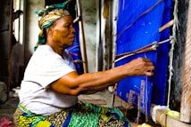 An elderly woman is sitting on the floor while weaving on a traditional loom. She is focused and uses her hands skillfully to manipulate the threads. She is wearing a colorful headscarf and a vibrant patterned skirt. The loom has blue fabric and several wooden components, situated in a modest room with muted wall colors.