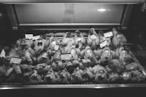 Rows of raw poultry are displayed inside a refrigerated glass case, each labeled with tags indicating price and type. The black and white image conveys a sense of a traditional butcher shop or market setting, emphasizing the variety of meats available for purchase.