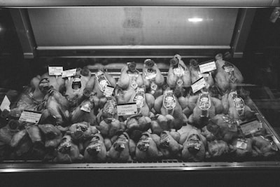 Rows of raw poultry are displayed inside a refrigerated glass case, each labeled with tags indicating price and type. The black and white image conveys a sense of a traditional butcher shop or market setting, emphasizing the variety of meats available for purchase.