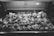 Rows of raw poultry are displayed inside a refrigerated glass case, each labeled with tags indicating price and type. The black and white image conveys a sense of a traditional butcher shop or market setting, emphasizing the variety of meats available for purchase.
