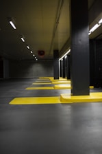 A dimly lit parking garage featuring multiple parking spaces marked by bright yellow lines. The concrete ceiling and floor have a gray hue, complemented by black support columns. Overhead fluorescent lights provide limited illumination, casting a subdued atmosphere.